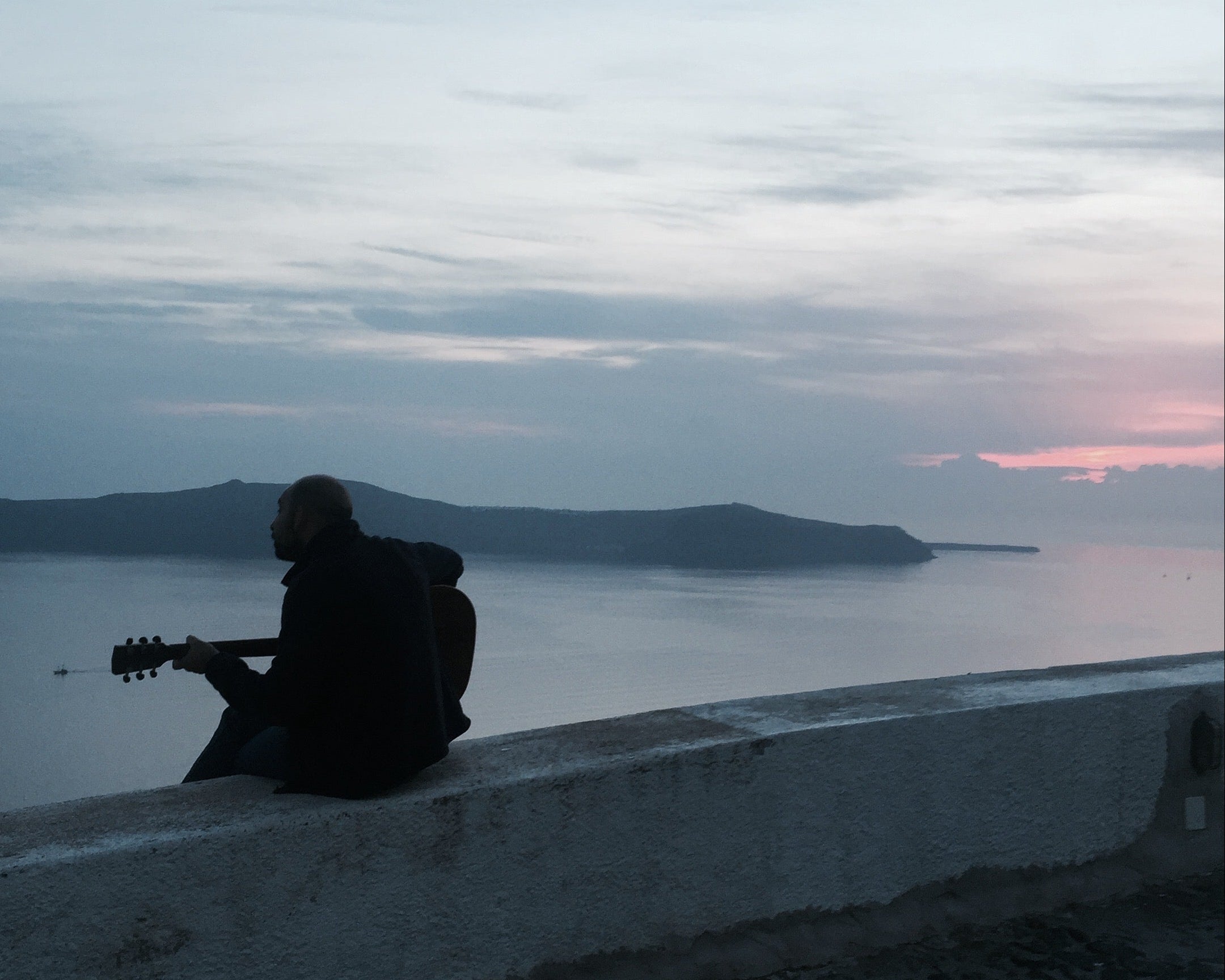Man playing guitar on the coast of Santorini, Greece at sunset, overlooking the Aegean Sea with golden light illuminating the shoreline.