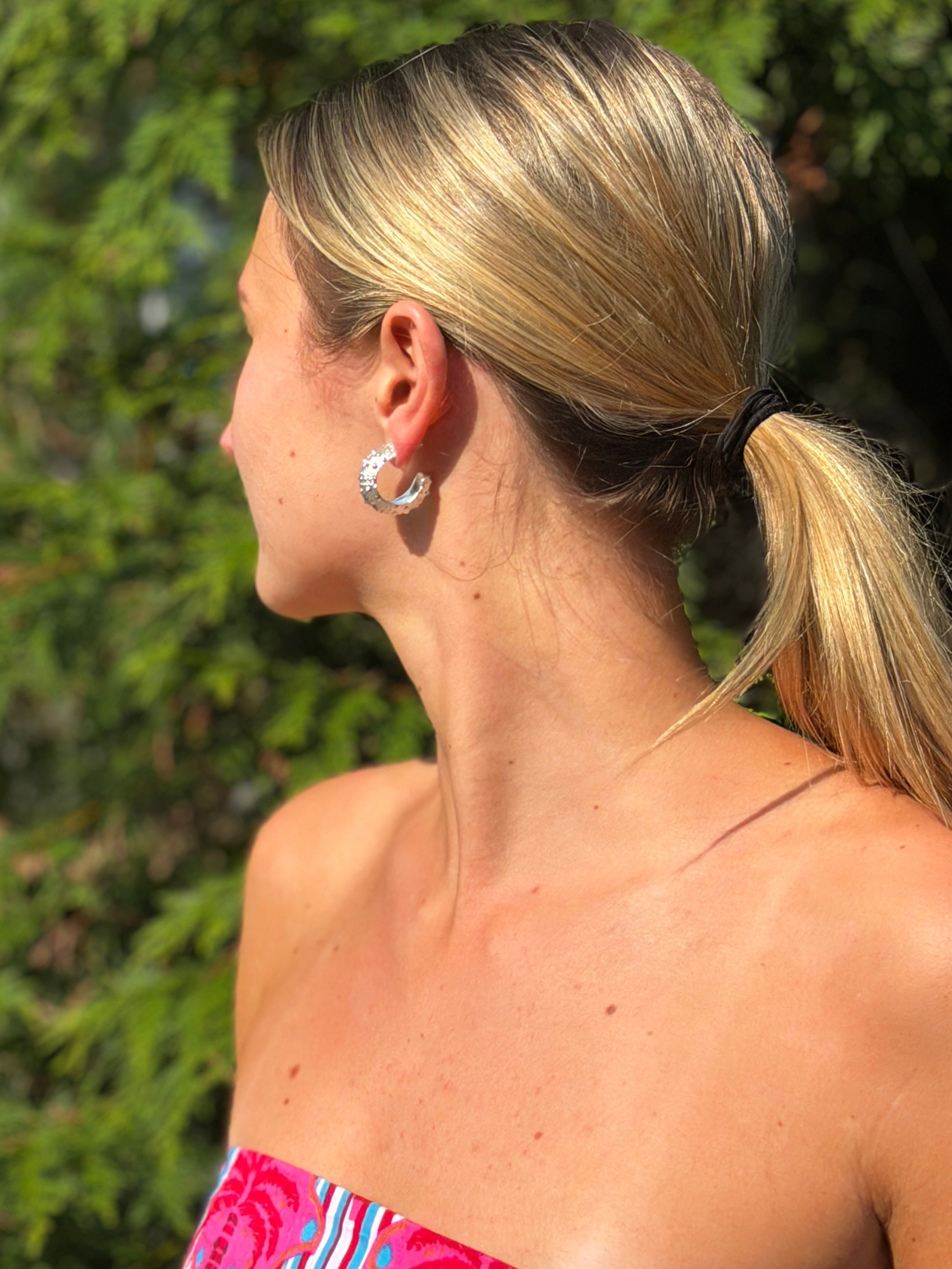 A woman in profile wearing a silver crescent-shaped hoop earring, photographed outdoors with green foliage in the background. sterling silver hoops made from sea urchin shells found while walking the beach.
