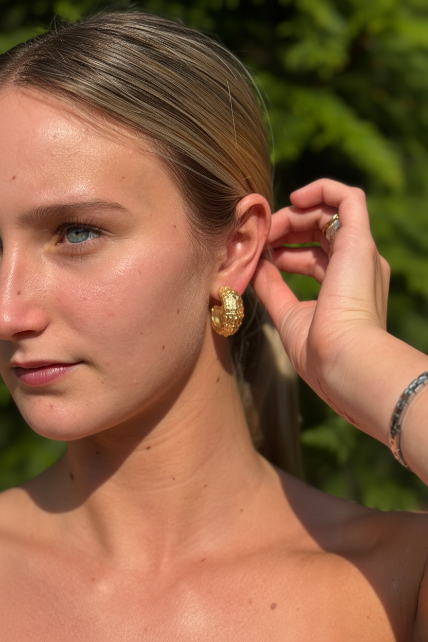 A close-up lifestyle shot of a woman wearing a textured gold hoop earring, with her hand adjusting the earring on her ear. The background is a soft-focus green outdoor foliage. Earrings made from sea urchin shells found while walking the beach in puerto rico